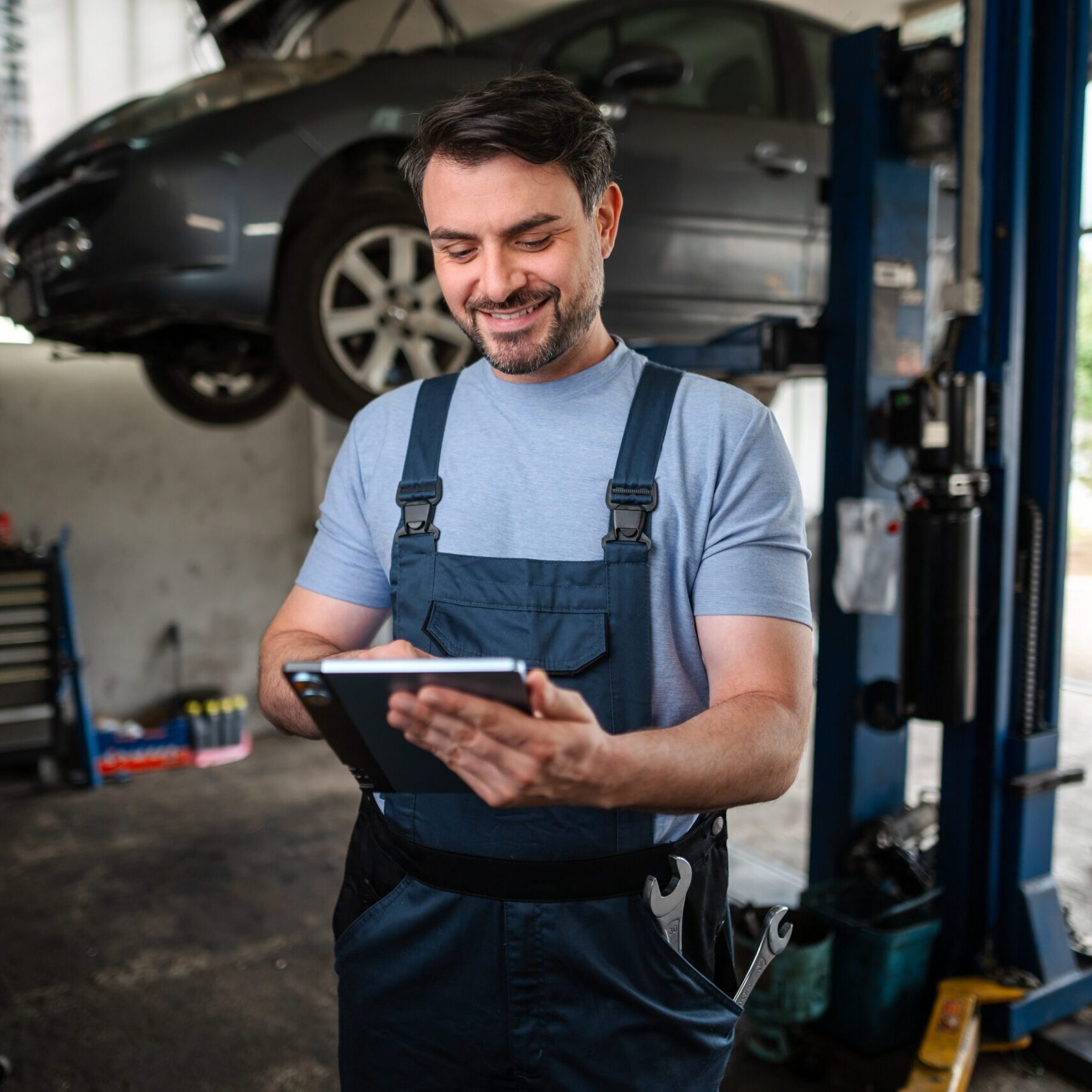 Smiling mechanic using a digital tablet next to a car on a hydraulic lift in a auto repair shop